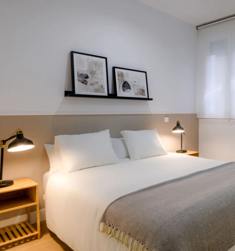 Un dormitorio moderno y minimalista con una cama de matrimonio, ropa de cama blanca, dos mesillas de noche con lámparas, una manta gris y dos cuadros de arte abstracto en una estantería.