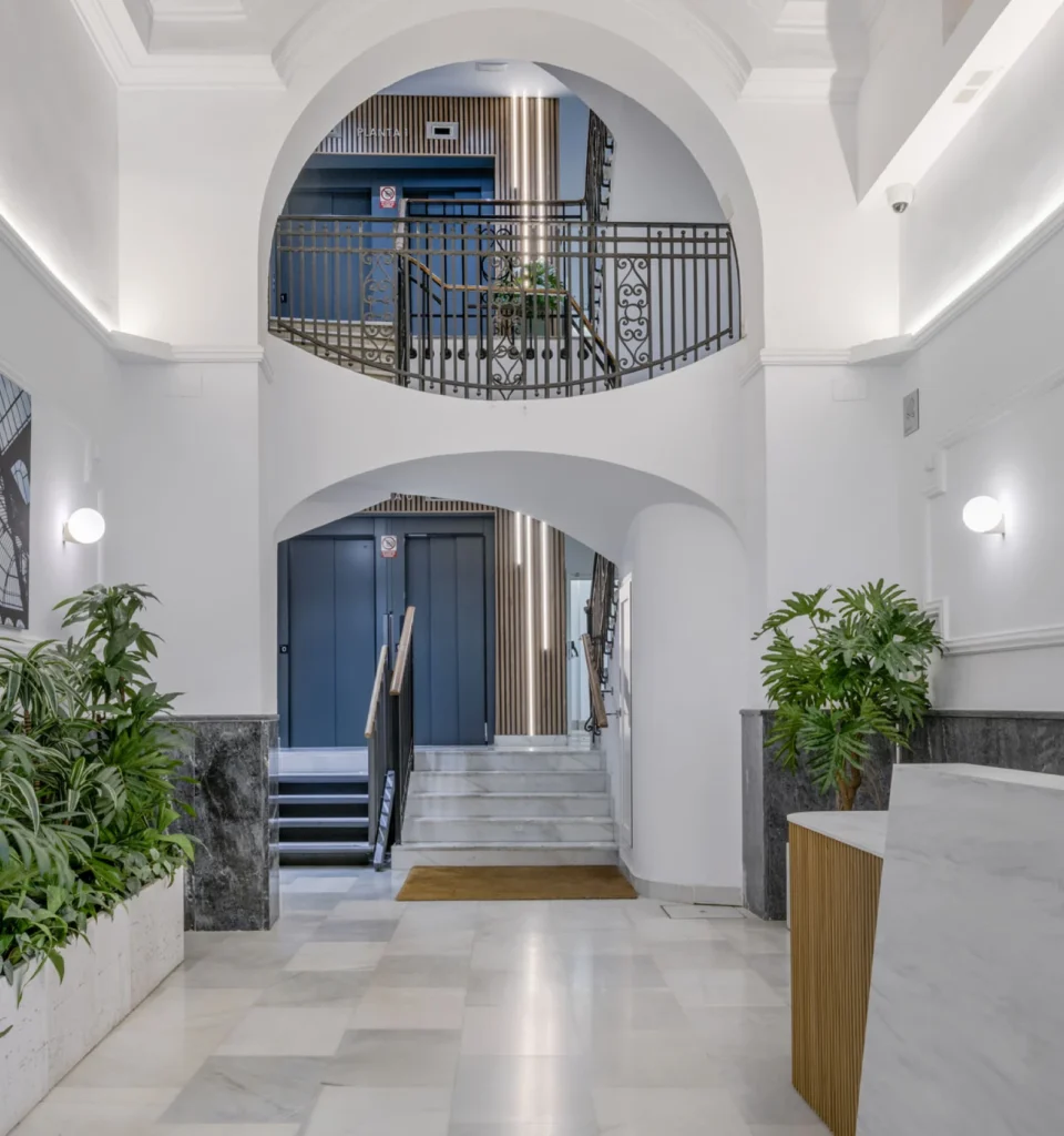 Vestíbulo de edificio moderno con arquitectura de arcos, suelos de mármol, plantas a lo largo de las paredes, una escalera que sube y una ornamentada barandilla metálica en el nivel superior. Una iluminación suave añade un ambiente acogedor.
