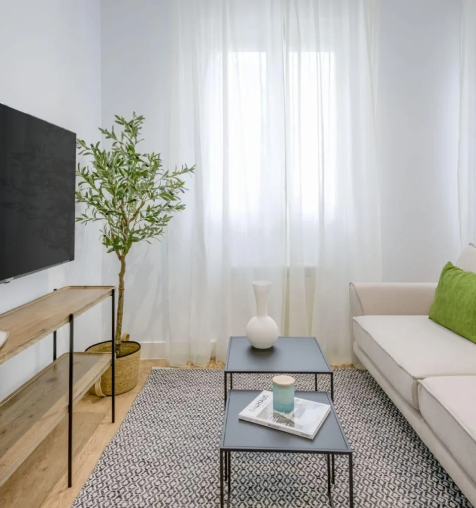 Un salón moderno con un sofá blanco y un cojín verde, una maceta, un televisor sobre un soporte de madera, dos mesas de centro de metal negro con decoración y cortinas blancas transparentes sobre una ventana.