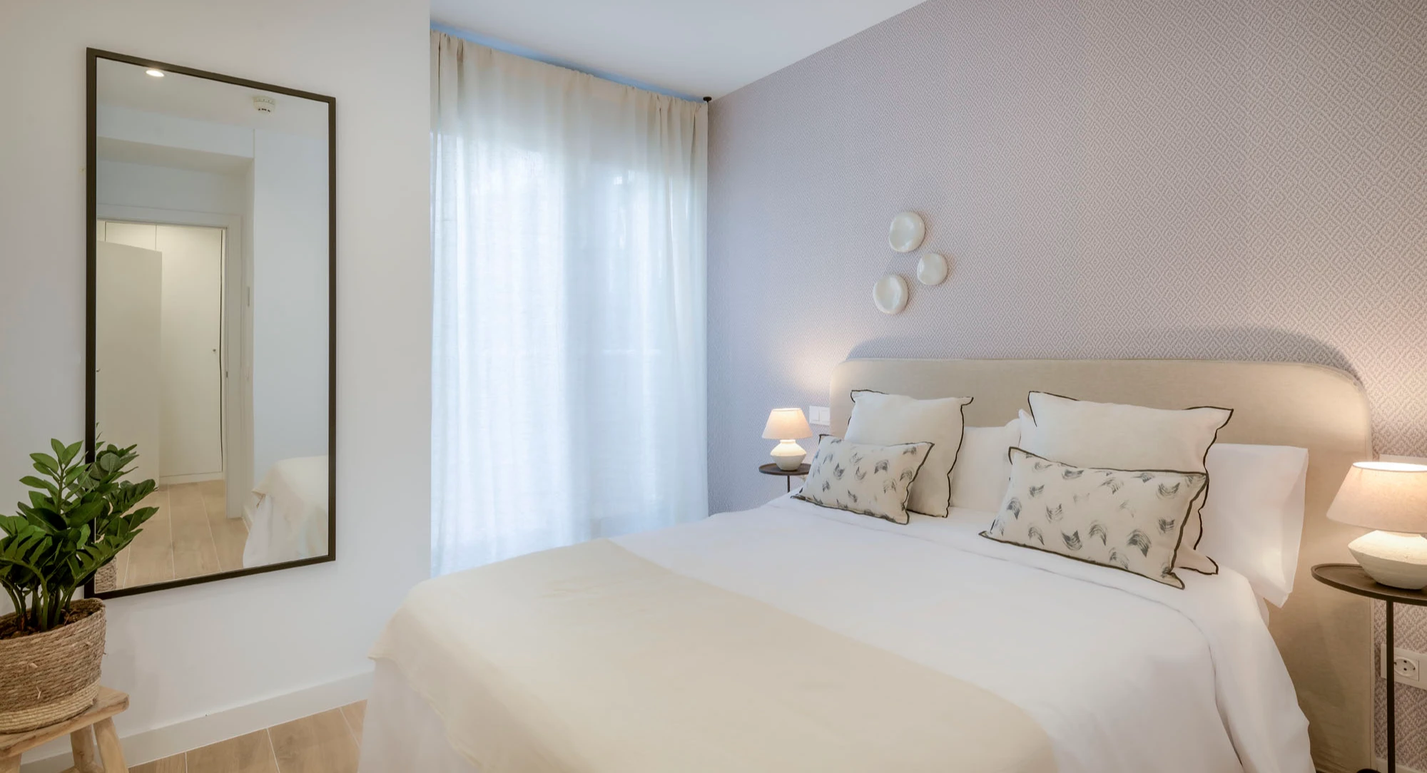 Un dormitorio moderno y acogedor con una cama doble, ropa de cama ligera, almohadas decorativas, dos mesillas de noche pequeñas con lámparas, arte mural sobre la cama, un gran espejo de pie y una maceta sobre el suelo de madera. La luz entra a raudales a través de cortinas transparentes.