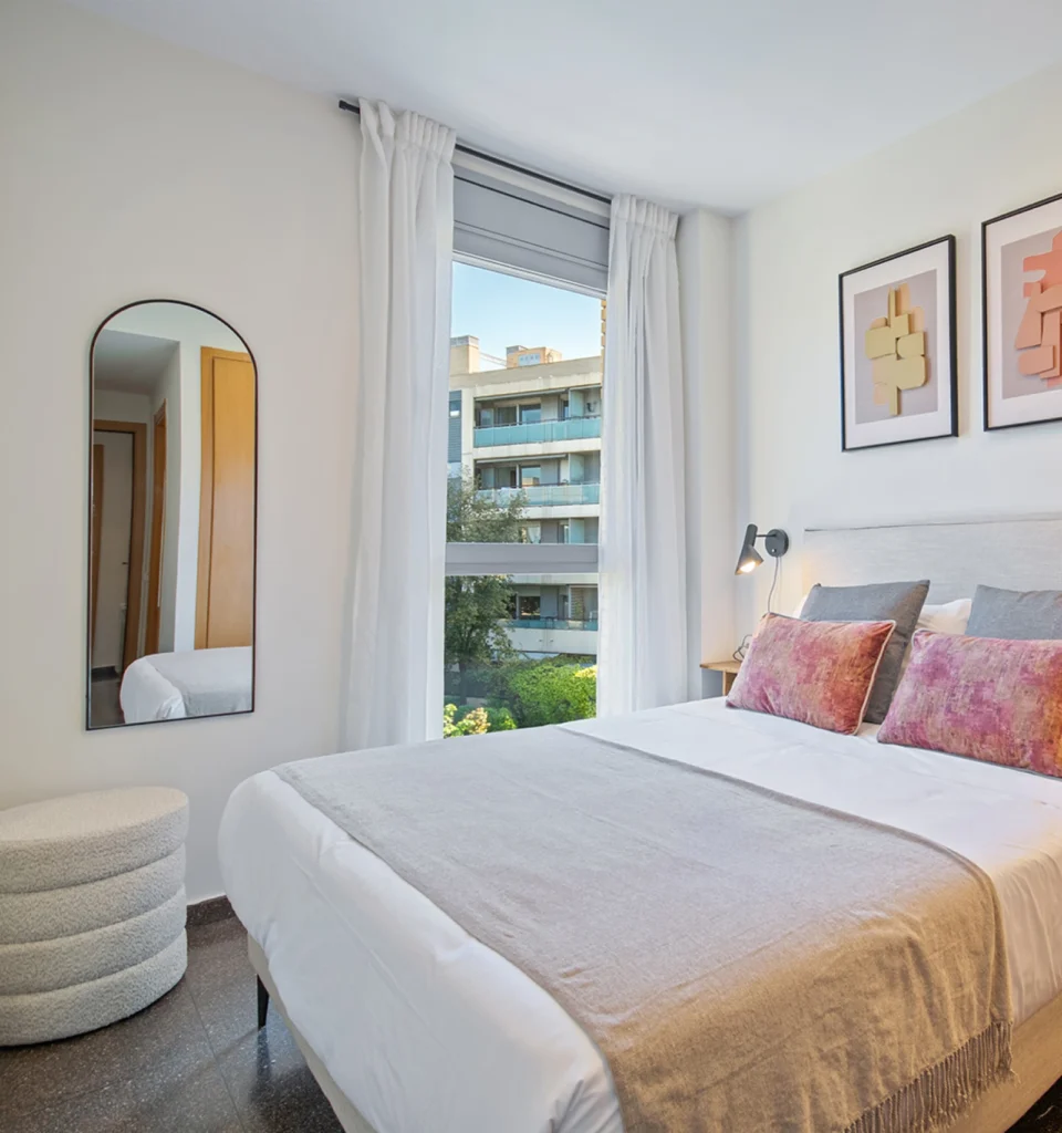 Un dormitorio con cama de matrimonio, ropa de cama blanca, cojines rosas y azules, una lámpara de pared, decoración sobre el cabecero, cortinas blancas hasta el suelo, un espejo de pared, una butaca redonda individual y una ventana con vistas a los modernos edificios del exterior.