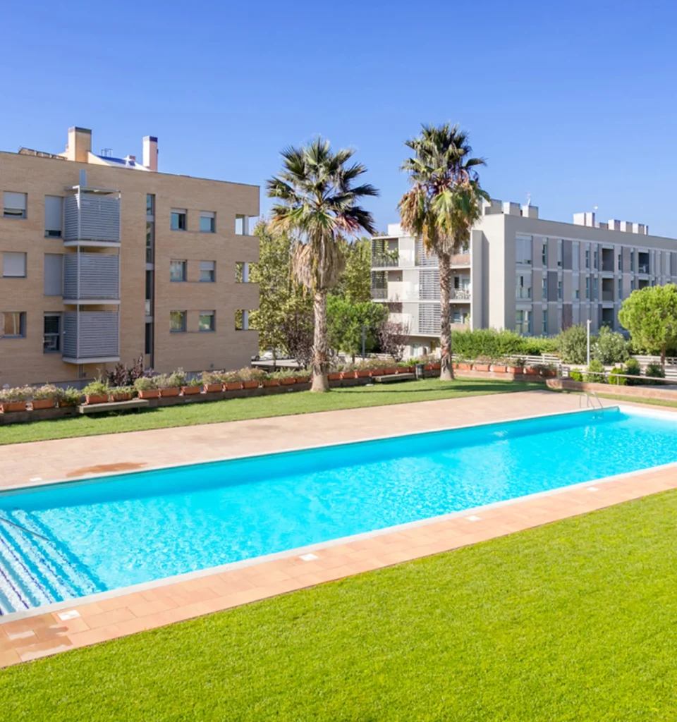 Una piscina rectangular al aire libre rodeada de césped verde y palmeras, con modernos edificios de apartamentos al fondo bajo un cielo brillante.