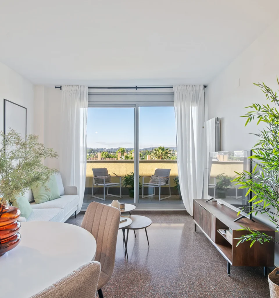 Sala de estar luminosa con un sofá de color blanco. Cuadros decorativos en la pared y un ventanal con vistas a un balcón con sillas y mesas. Cortinas blancas hasta el suelo y un aparador de madera sosteniendo un televisor.