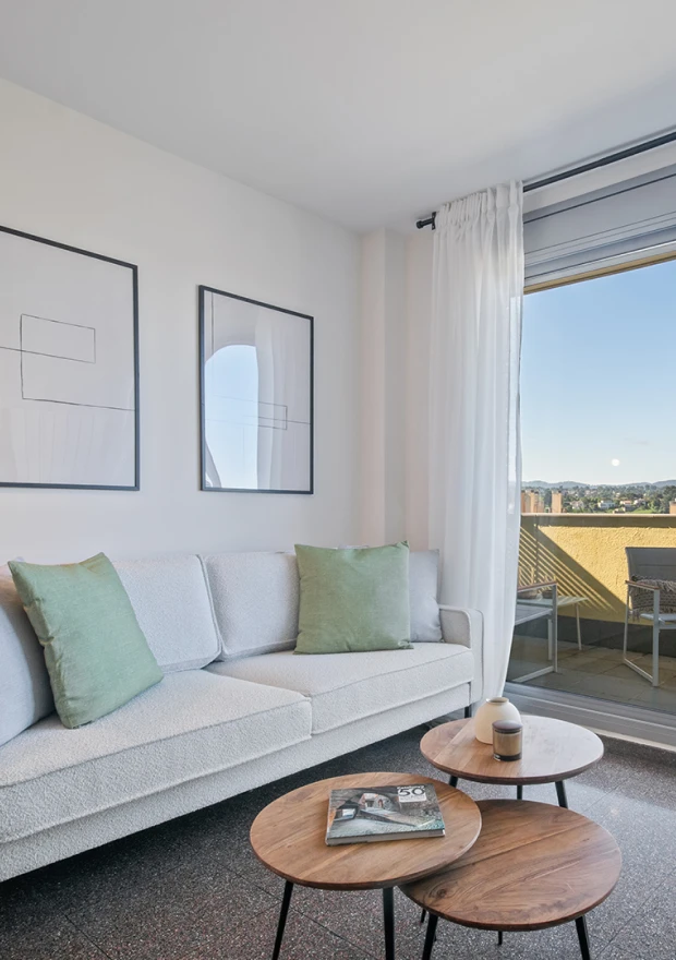Moderno salón con sofá blanco, cojines verdes, dos mesas de centro redondas de madera y cuadros abstractos en la pared. Una gran ventana con cortinas transparentes da a un balcón con vistas panorámicas al exterior.