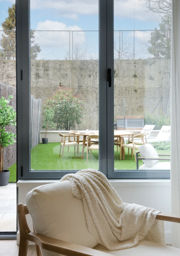 Un acogedor sillón crema con una manta texturizada está sentado junto a una gran ventana, con vistas a un patio verde con una mesa, sillas y plantas contra un muro de piedra bajo un cielo azul con nubes.