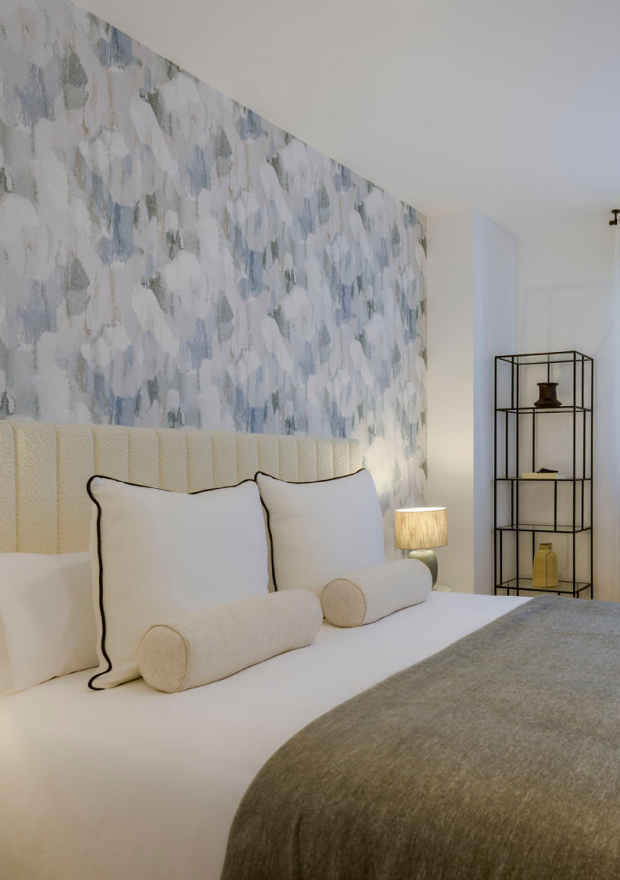 Un dormitorio moderno con una cama con sábanas blancas, dos almohadas grandes y dos cojines cilíndricos. Detrás de la cama hay una pared de acento estampada y en la esquina una estantería de metal negro con adornos. Sobre la mesilla de noche hay una lámpara.