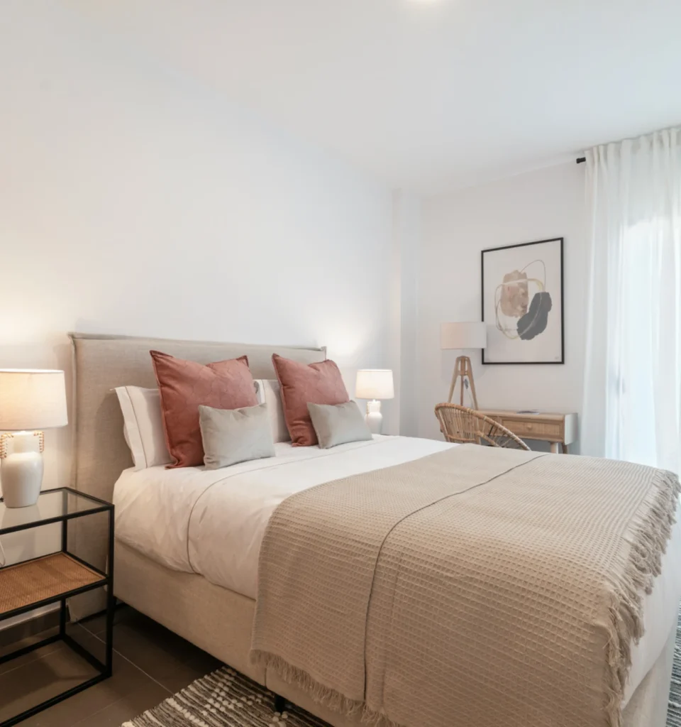 Un dormitorio con una cama de matrimonio, con ropa de cama de color beige y blanca. Dos mesillas de noche con lámparas, cojines rosas. Una ventana con cortinas blancas.