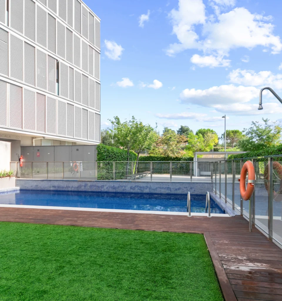 Una piscina al aire libre junto en el exterior de una urbanización residencial, con una valla de cristal, una ducha exterior y césped artificial.