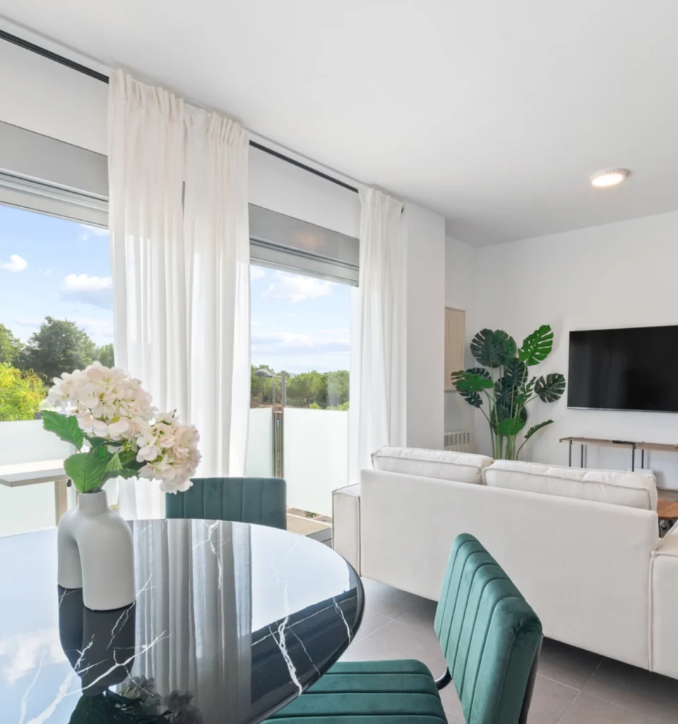 Un moderno salón con una mesa de comedor redonda de mármol negro, sillas de terciopelo verde, sofá blanco, televisor en la pared, una planta alta y grandes ventanales con cortinas blancas.