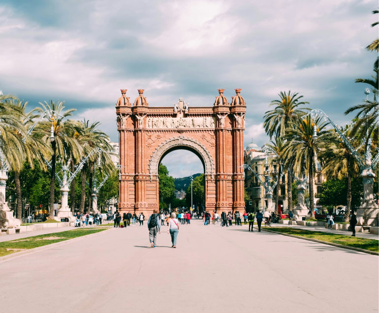 Un amplio paseo conduce al Arco del Triunfo, monumento de ladrillo rojo de Barcelona, bordeado de palmeras y gente paseando bajo un cielo parcialmente nublado.