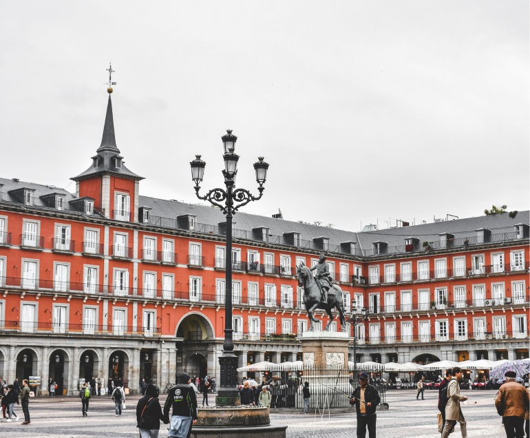 Gente paseando por la Plaza Mayor de Madrid, con una farola central y una estatua ecuestre. Edificios históricos rojos con muchas ventanas y tejados grises rodean la plaza en un día nublado.