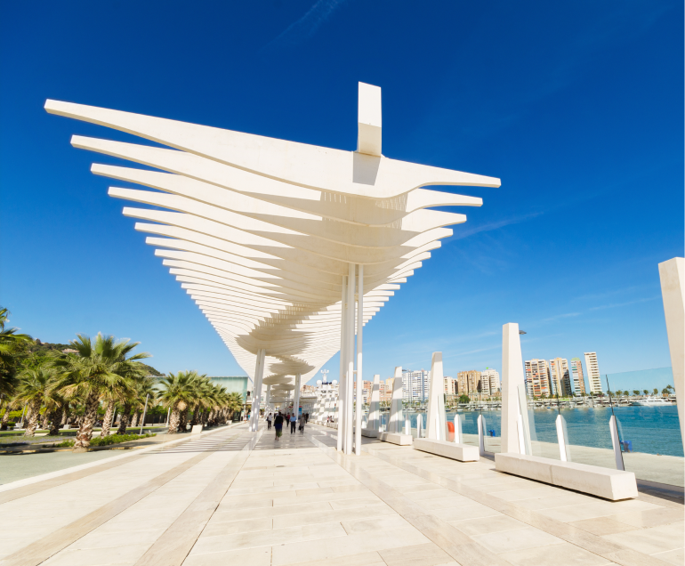Una moderna pérgola blanca con artísticas vigas en un paseo marítimo. Rodeado de palmeras y gente paseando.
