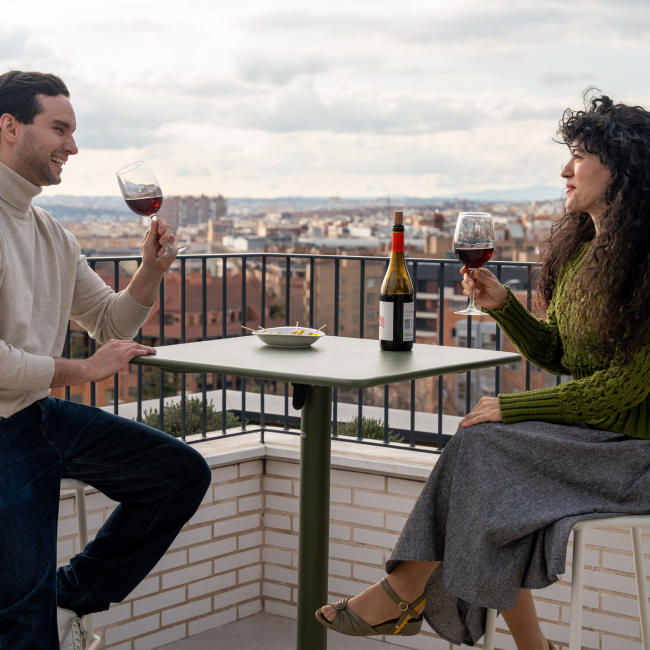 Un hombre y una mujer se sientan de frenteen una mesa en la azotea, con vasos de vino tinto. Sobre la mesa hay una botella de vino y un pequeño plato. Al fondo, edificios urbanos.