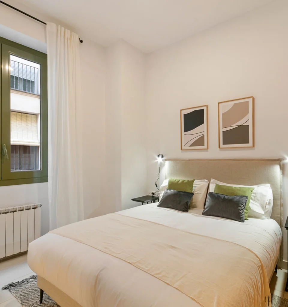 Un acogedor dormitorio con cama de matrimonio, ropa de cama clara, cojines de acento verdes y grises, dos obras de arte abstracto sobre el cabecero, una lámpara de mesilla y una ventana con cortinas blancas que deja pasar la luz natural.