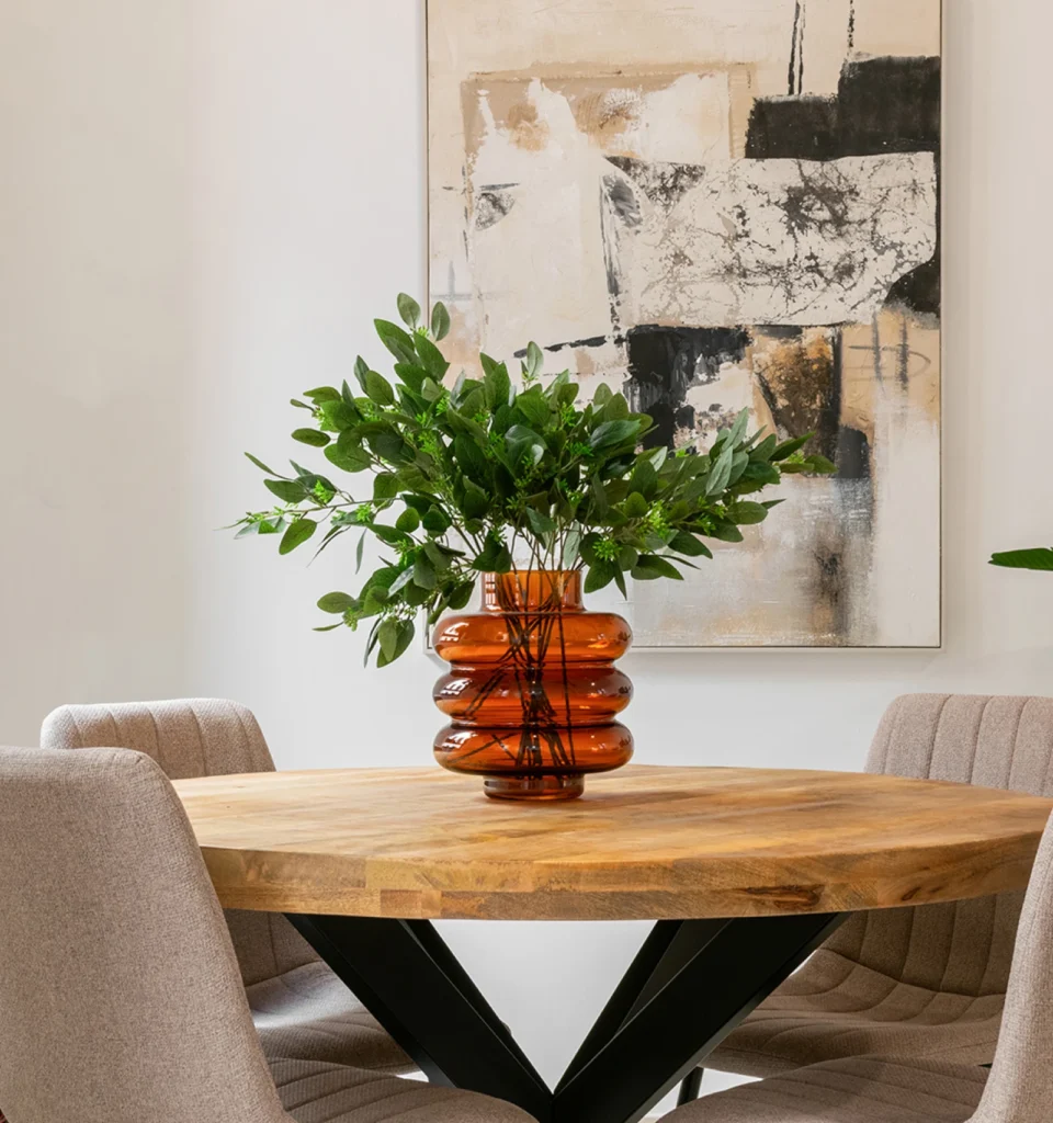 Una mesa redonda de madera con un jarrón de cristal marrón con ramas de hojas verdes. Cuatro sillas acolchadas de color beige y paredes neutras. Un cuadro decorativo en la pared.