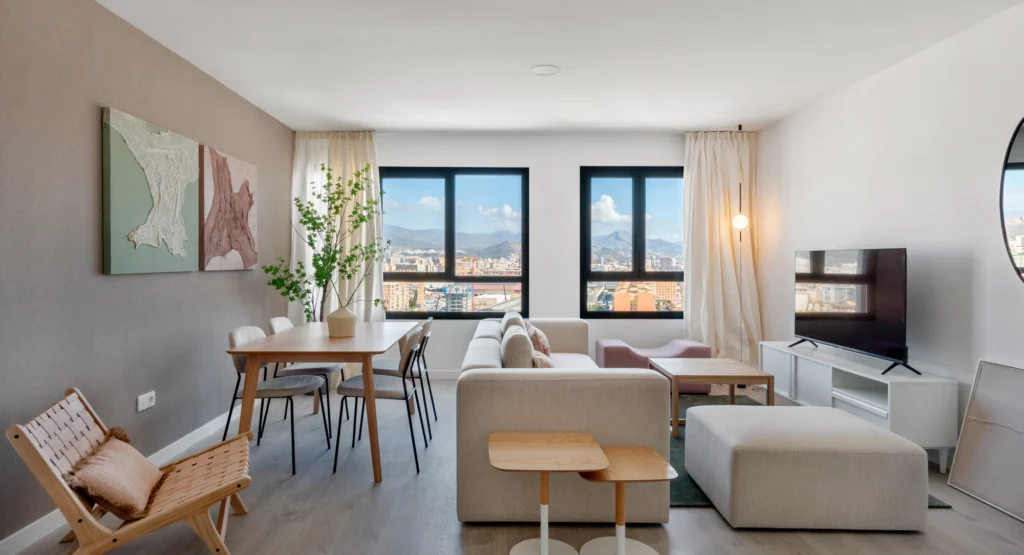 Moderno salón-comedor con muebles de madera clara, sofá beige, grandes ventanales que muestran un paisaje urbano con montañas, decoración neutra, un televisor y una obra de arte en la pared. El salón es luminoso y está ordenado.