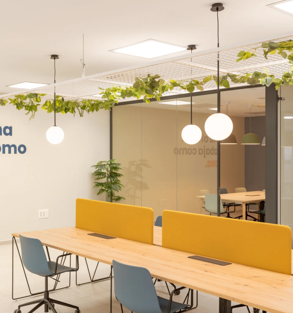 Una oficina moderna con mesas de madera, separadores amarillos, sillas azules, lámparas redondas colgantes, vegetación en el techo, una maceta y una sala de reuniones con paredes de cristal al fondo.