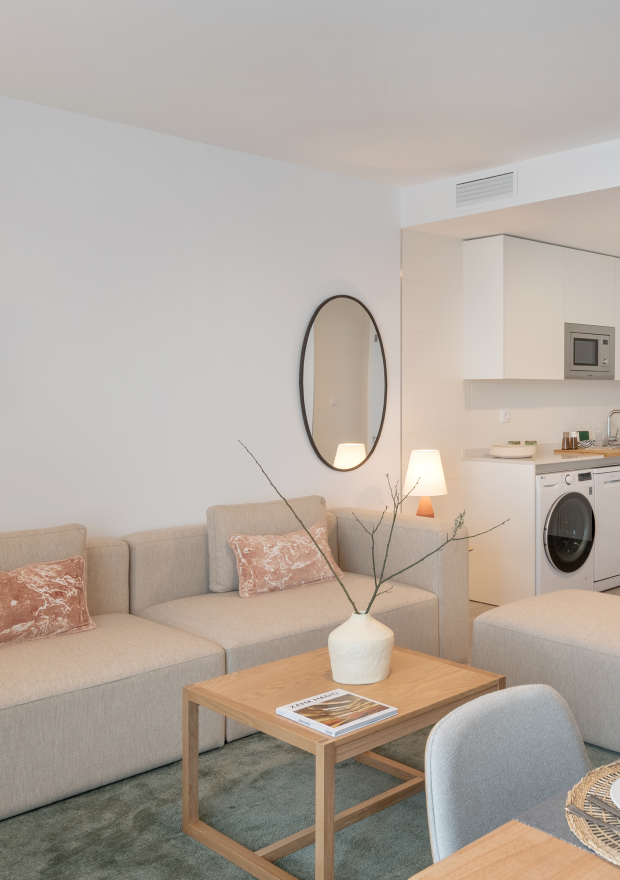 Un moderno salón con un sofá beige, cojines estampados, una mesa de centro de madera con un jarrón y revistas, un espejo de pared redondo y una vista de una zona de cocina blanca con lavadora y electrodomésticos.