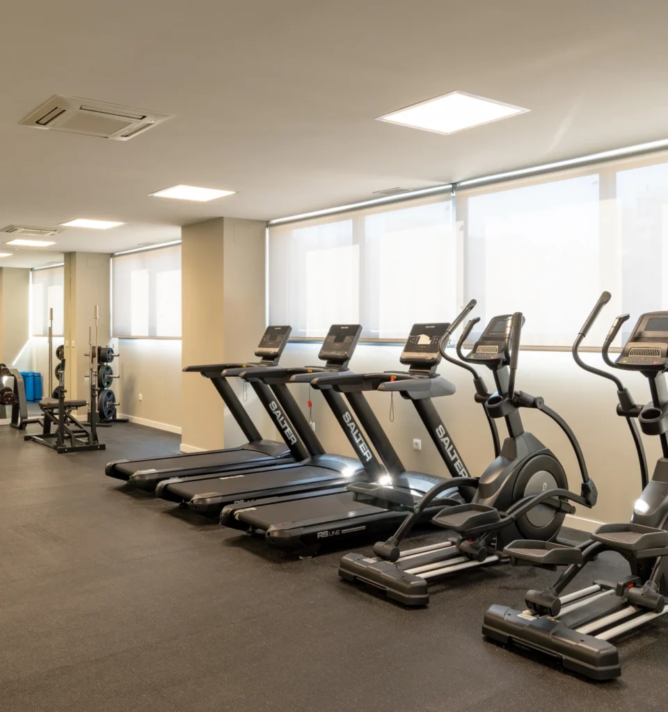 Un gimnasio moderno con cintas de correr y máquinas elípticas alineadas cerca de grandes ventanales sobre un suelo de caucho. El espacio está iluminado con luz natural y artificial.