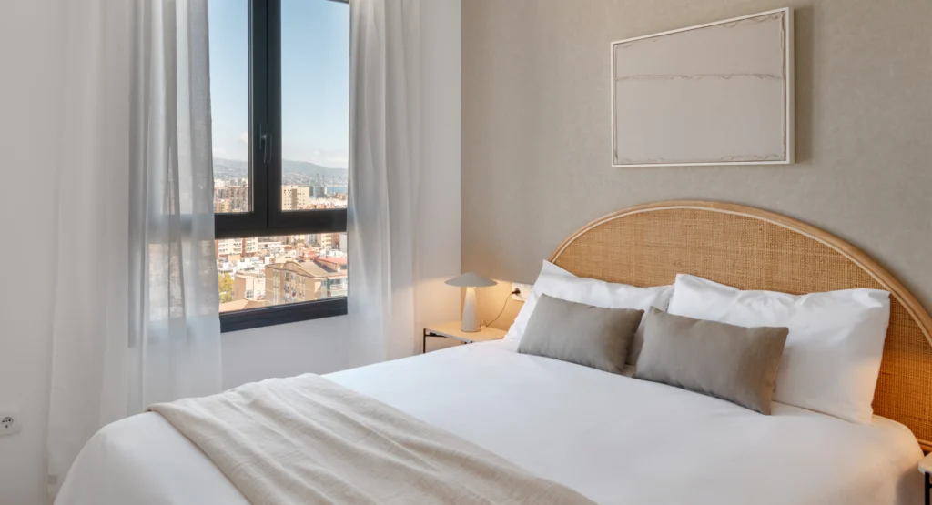 Dormitorio luminoso y moderno con una cama grande, sábanas blancas, almohadas neutras, un cabecero de ratán, una ventana con vistas a la ciudad, cortinas transparentes, una lámpara de noche y arte minimalista en la pared.
