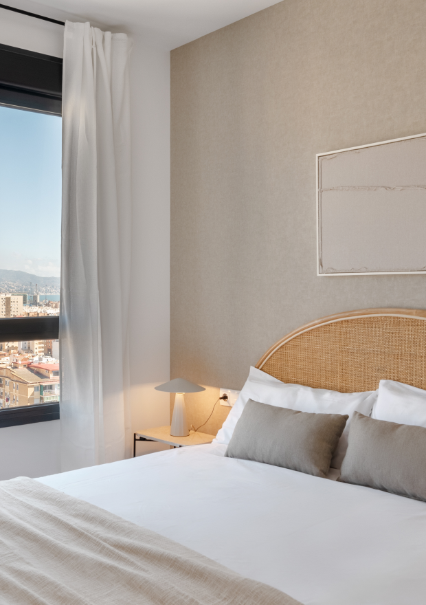 Un dormitorio luminoso y moderno con una cama grande de matrimonio, ropa de cama blanca, dos almohadas grises, un cabecero de mimbre, una mesita de noche de madera con una lámpara, una ventana con cortinas blancas y vistas a la ciudad.