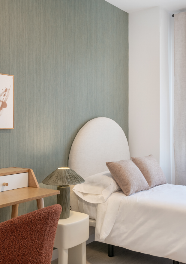Un dormitorio con una cama individual, dos almohadas de color beige, un cabecero blanco redondeado, una mesita auxiliar con una lámpara, un escritorio de madera y una silla de color granate contra una pared de color verde claro.
