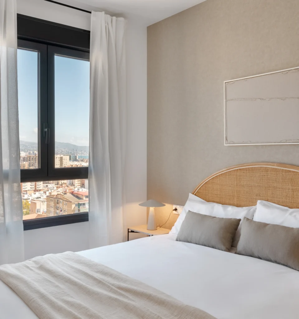 Un dormitorio moderno con un ventanal con vistas a la ciudad. La habitación cuenta con una cama individual con sábanas blancas, una manta y cojines beige, un cabecero de mimbre. Mesitas de noche con una lámpara y paredes de colores neutros.