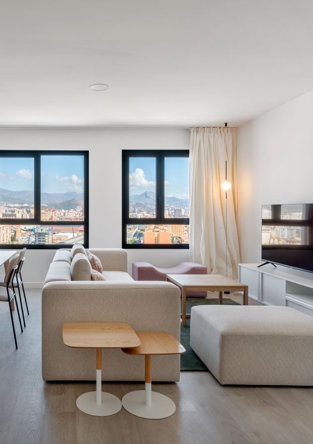 Moderna sala de estar con sofá seccional beige claro, otomana, mesas auxiliares de madera, TV y grandes ventanales con vistas a la ciudad y las montañas a lo lejos. En la esquina con cortinas hay una lámpara de pie.