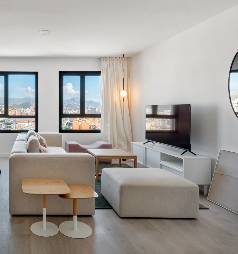 Un salón con suelo de madera clara, un sofá beige, mesitas de madera, un televisor sobre un mueble blanco, grandes ventanales con vistas a la ciudad y cortinas transparentes. También se ven un espejo redondo y una lámpara de pie.