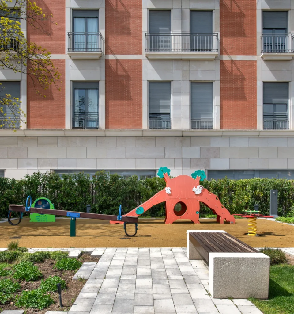 Pequeño parque infantil con forma de dos animales naranjas. Cerca hay bancos y zonas verdes. Al fondo, la fachada de un edificio de ladrillos rojos y blancos.