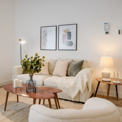 Un salón moderno con un sofá crema, cojines neutros y una manta beige. Una mesa de centro de madera sostiene un jarrón con plantas y velas. Dos obras de arte abstracto cuelgan de la pared sobre el sofá.