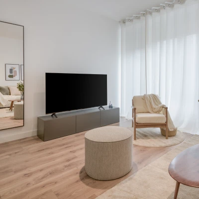 Un salón moderno y minimalista con un gran espejo, un televisor de pantalla plana sobre una consola baja, una otomana redonda, un sillón acolchado con una manta, suelo de madera clara y cortinas blancas.