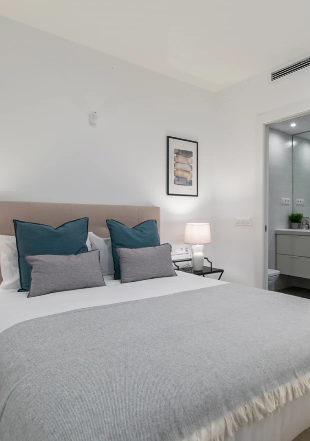 Un dormitorio moderno y minimalista con una cama bien hecha con almohadas azules y grises, una manta gris, una mesilla de noche con una lámpara, obras de arte en la pared y vistas a un luminoso cuarto de baño.