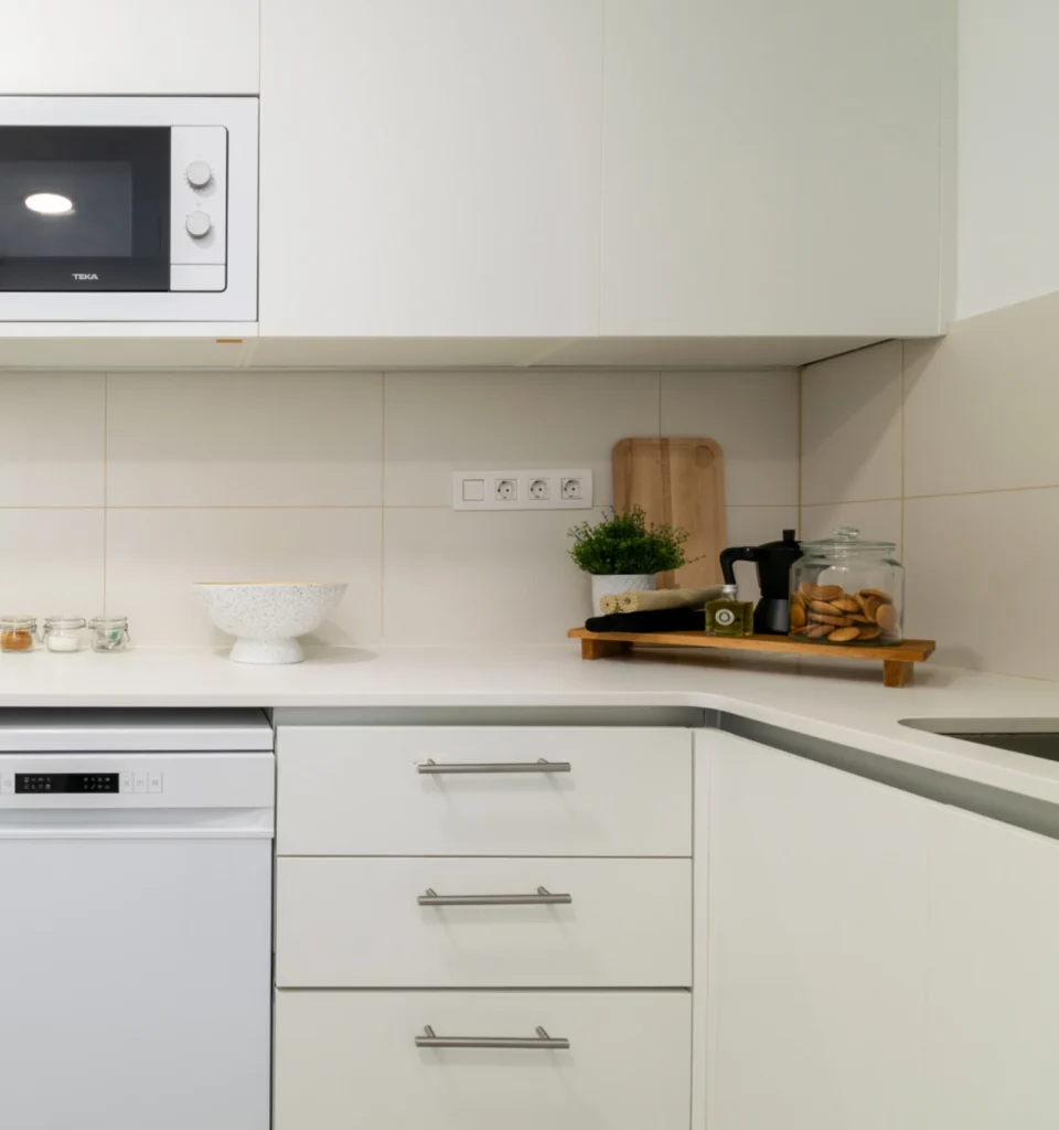 Cocina moderna con muebles de color blanco, equipada con lavavajillas, microondas y fregadero. Sobre la encimera, hay una tabla de cortar y algunos recipientes.
