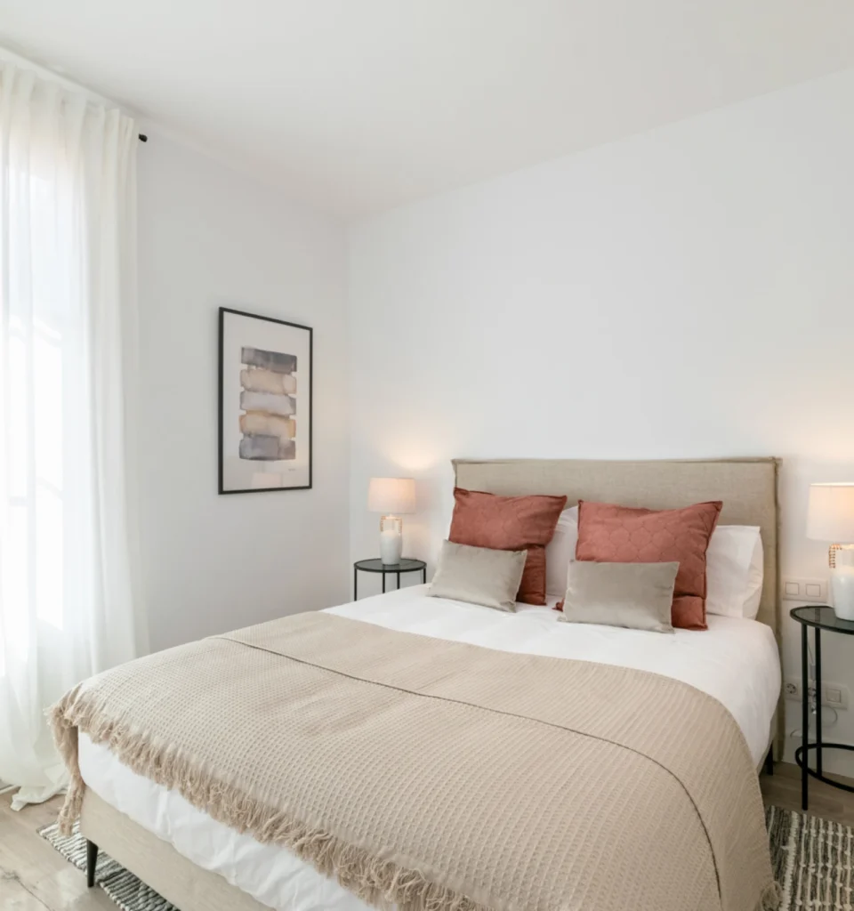 Un dormitorio luminoso con una cama de matrimonio, almohadas grises y de color granate, dos mesillas de noche con lámparas pequeñas, una obra decorativa en la pared, cortinas blancas transparentes y suelo de madera clara.