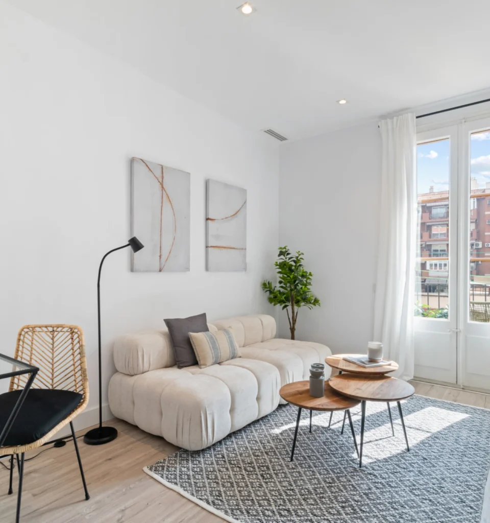 Luminoso salón moderno con un sofá beige, dos mesas de centro redondas de madera, una alfombra estampada, una lámpara de pie, una maceta y arte en la pared. Grandes ventanales con cortinas blancas.
