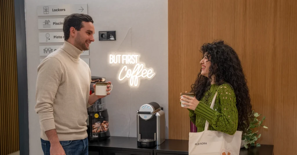 alt="Dos residentes charlando en la zona común con café, junto a máquina de café y cartelería de servicios del edificio"