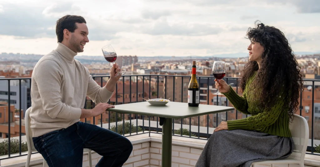 Un hombre y una mujer se sientan en una mesa en la azotea, sonrientes y con copas de vino tinto en la mano. Sobre la mesa hay una botella de vino y un pequeño plato de comida. Al fondo se ven edificios urbanos y un cielo nublado.